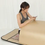Woman half-sitting inside infrared sauna dome using smartphone, flexible multi-position home far infrared therapy pod